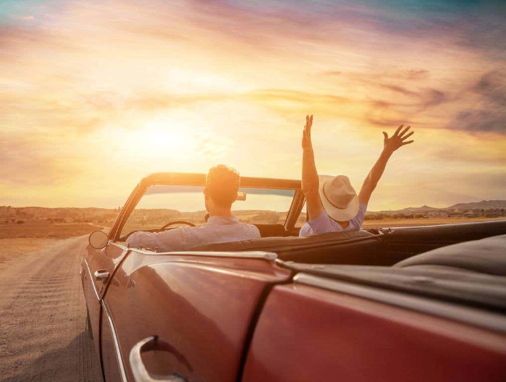 Photo of a man and woman who love taking road trips. The couple are riding in a vintage convertible car with the top down. The man is driving. The woman in the passenger seat is wearing a straw cowboy hat and has her hands and arms raised above the front windshield signifying sheer joy and happiness. The car is heading down an empty two-lane highway towards a golden sunset on the horizon.