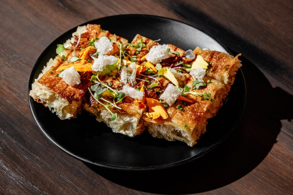 A black plate on a wooden table holds sliced bread topped with various Italian toppings.