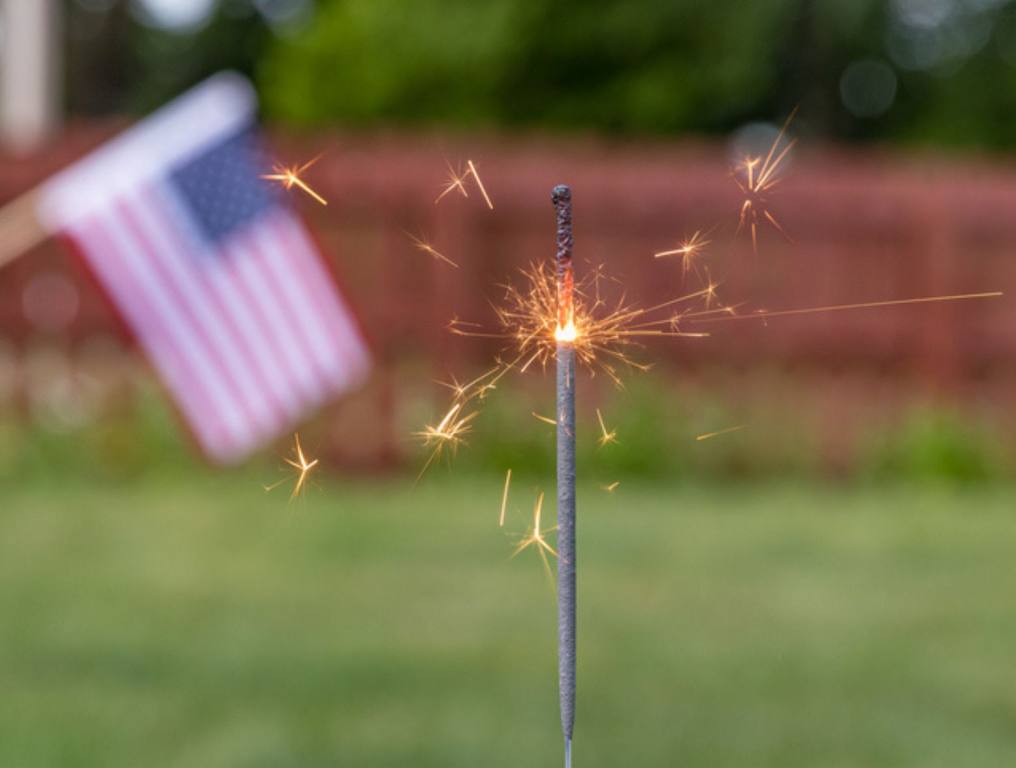 Closeup of burning sparkler and American flag in backyard. Concept of fireworks safety, Independence Day and Fourth of July celebration