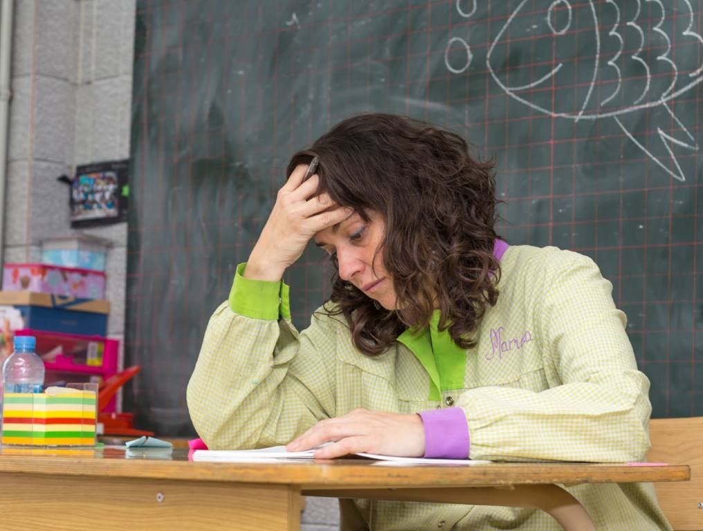 Industries with highest burnout rates include the education sector. Photo of a grade school teacher sitting at her classroom desk. She is leaning her head onto her right hand and arm. She is staring at the paper on her desk. She is sitting in front of a black chalkboard with chalk writing on it. There is a pad of colorful sticky notes to her right on the desktop.