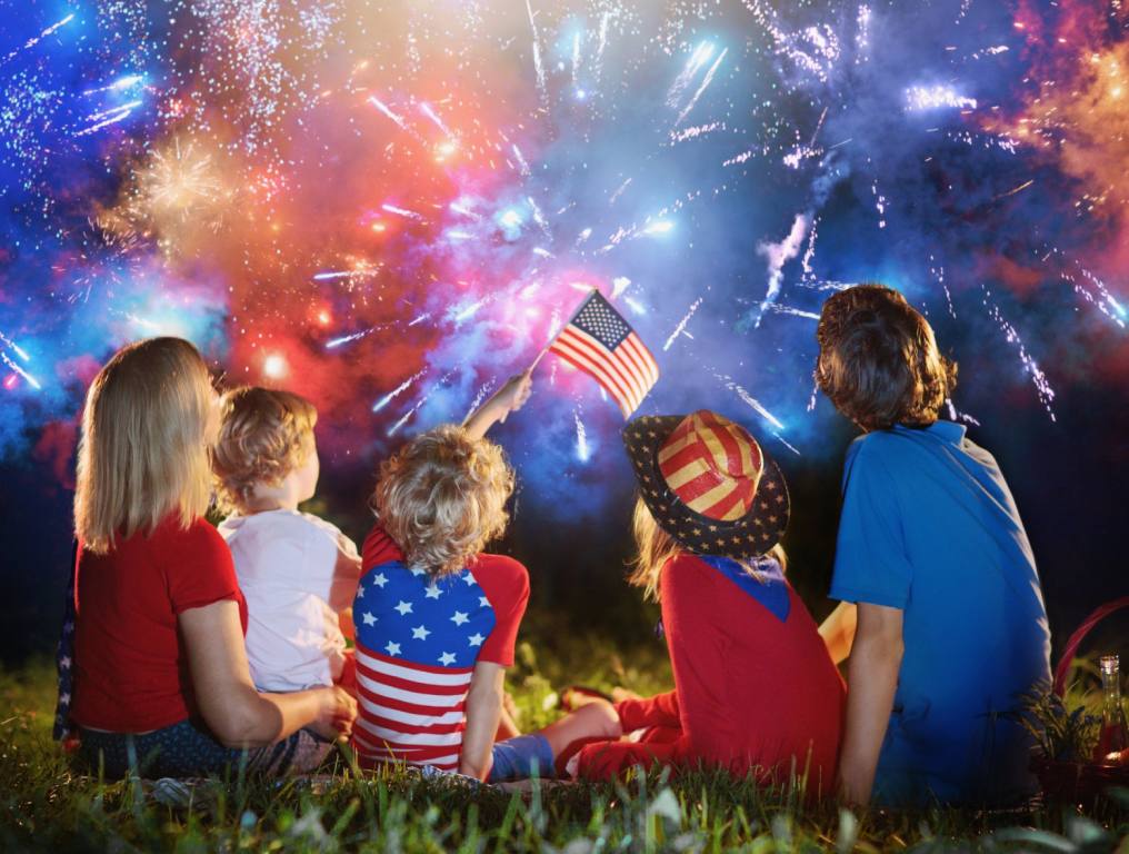 A behind shot of a family of five sitting on a lawn and wearing patriotic colors and watching 4th of July fireworks while waving small American flags.