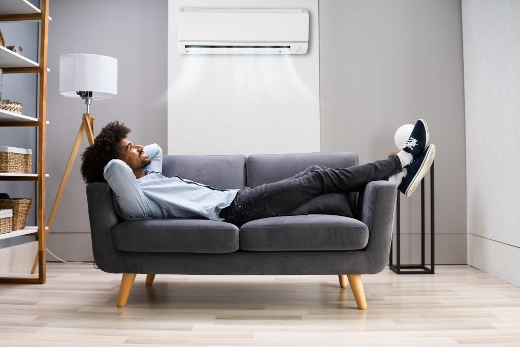 A young black man lays on a modern gray loveseat with his hands behind his head and his feet propped up as he enjoys cool air coming from a wall unit above him.