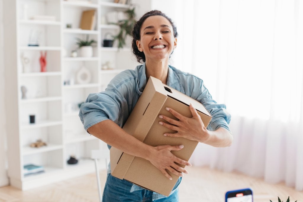 Joyful Middle Eastern Lady Holding Cardboard Box Receiving Delivered Package Standing At Home, Smiling With Eyes Closed.