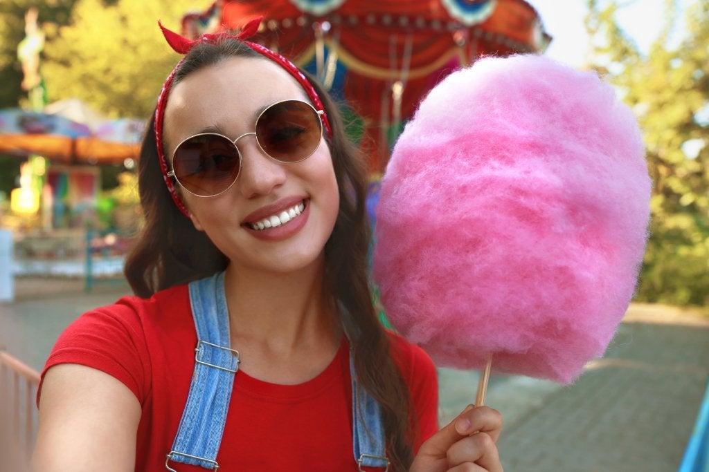 A young girl in sunglasses and a red shirt and denim overalls smiles at the camera with a pink cotton candy in her hand.