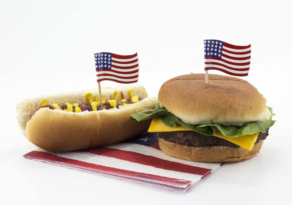 A hot dog and hamburger sit on a white table atop an American flag napkin with American flag toothpicks stuck in them.