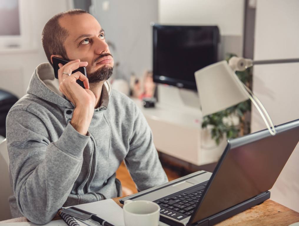 A balding white man in a gray hoodie is on the phone looking frustrated while he sits in front of an open laptop. Concept: Lost Online Orders