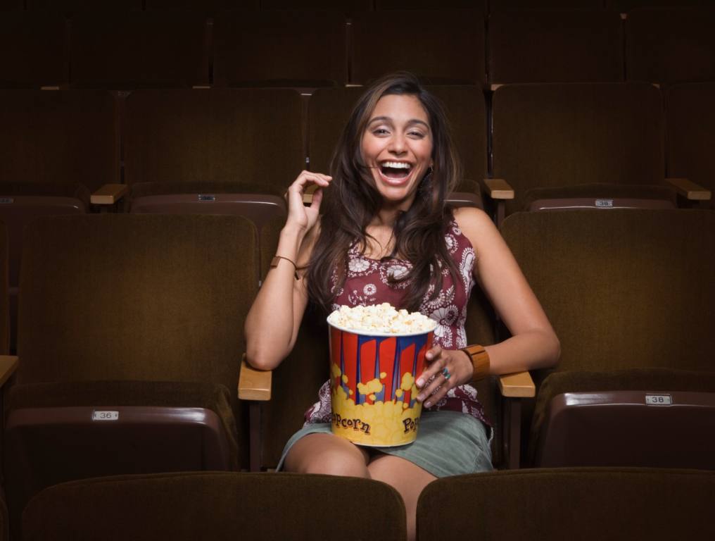 A young lady sitting at the movies by herself and having a good time with some popcorn.