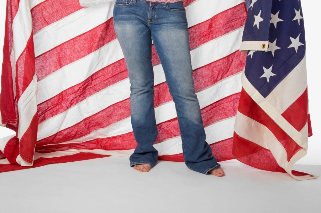 The lower half of a woman in blue jeans holding a giant American flag behind her.