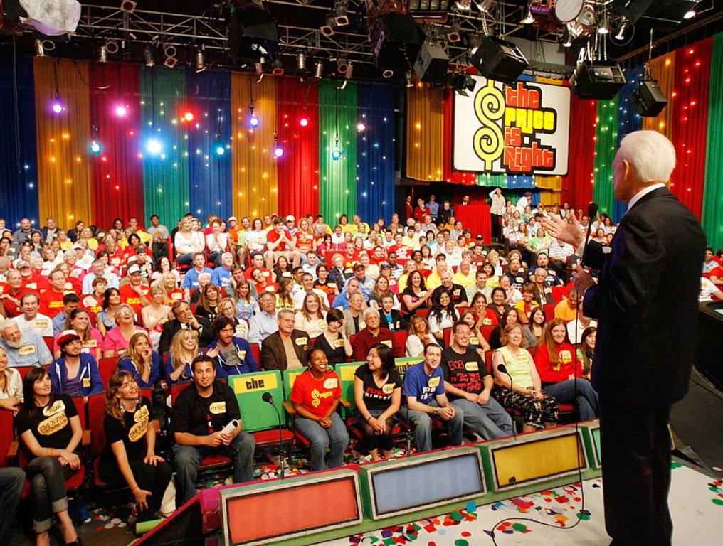 The Price Is Right is coming to Henderson. Photo of legendary host of that TV game show at his final taping in 2007. Bob Barker back and left side are visible as he addresses the audience. He is wearing a dark suit and his hair is white. He is holding a microphone in his left hand and gesturing with his right hand. "The Price Is Right" sign can be seen in the back right side of the studio. The audience is packed with people wearing colorful clothing. The podiums where new contestants stand to make their first bids can be seen at the foot of the stage upon which Bob Barker is standing.