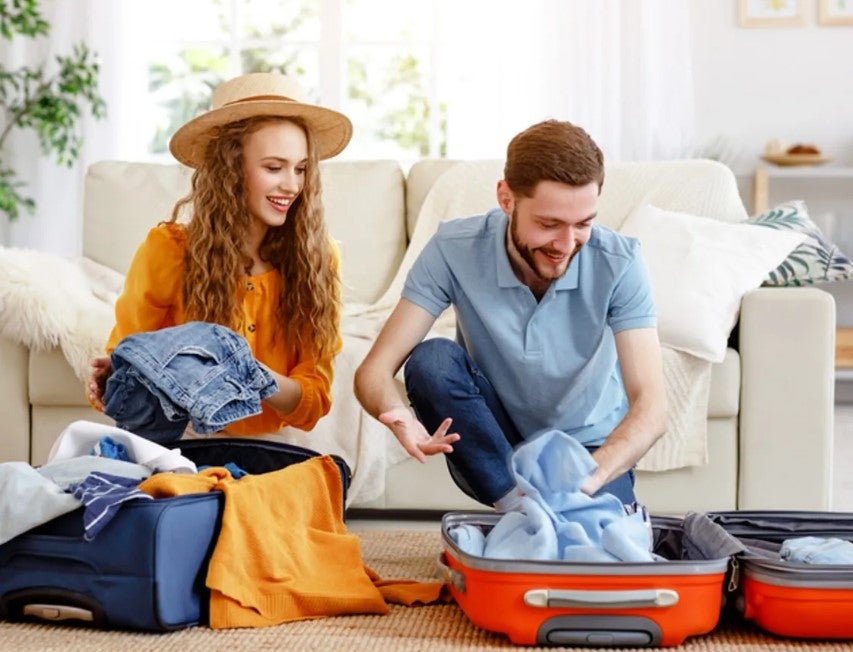 Laughing man and woman in summer hat setting off for journey and packing bags on floor in living room anticipating vacation, pre-vacation prep concept