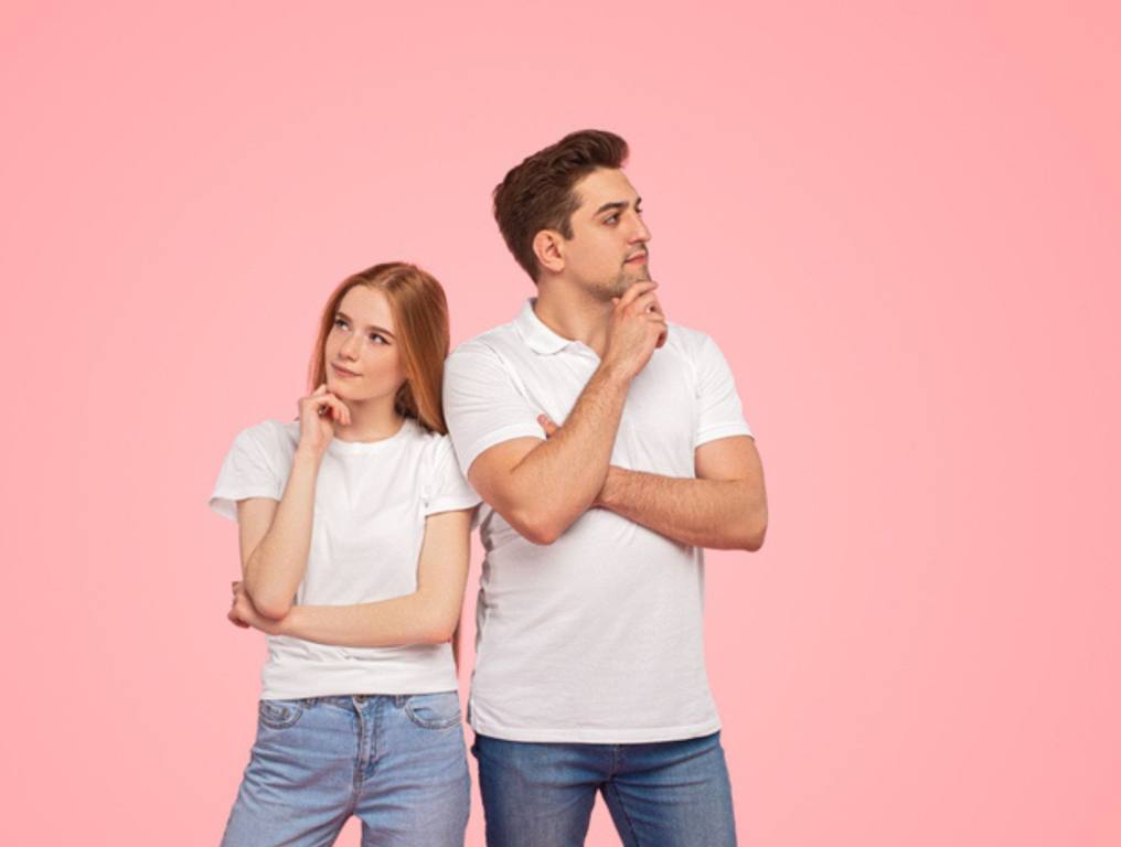 Pensive young man and woman in similar clothes touching chins and thinking while standing near each other and looking in different directions against pink background