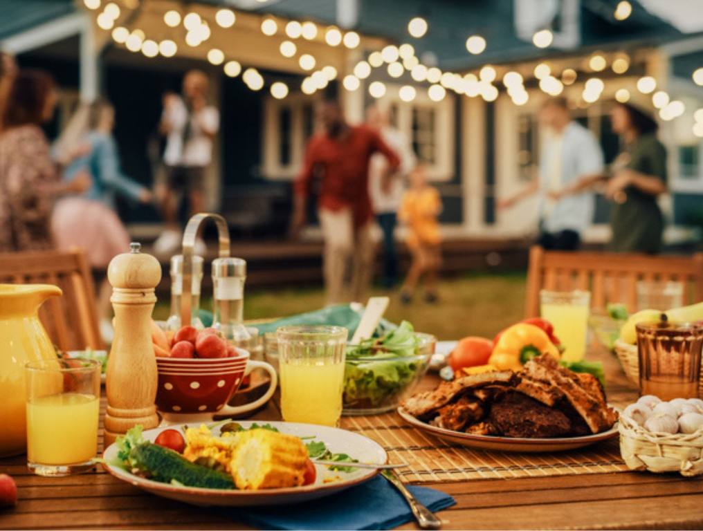 Backyard Dinner Table with Tasty Grilled Barbecue Meat, Fresh Vegetables and Salads. Happy Joyful People Dancing to Music, Celebrating and Having Fun in the Background on House Porch. Summer Cookout Concept