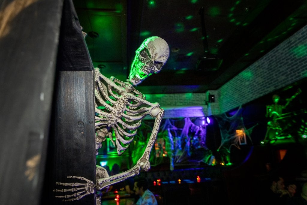 A smiling skeleton Halloween decoration is peaking out from behind a wall in a Halloween-themed bar.