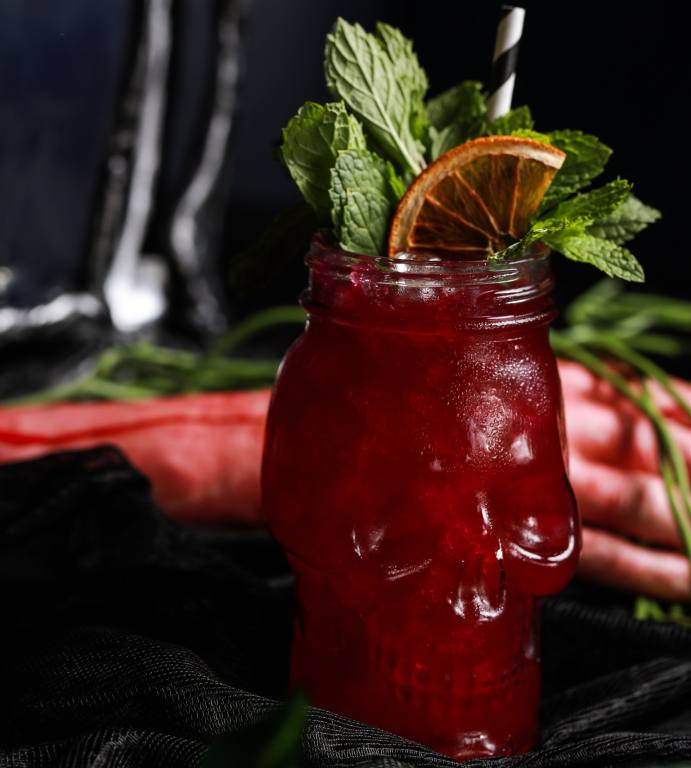 A cocktail in a skull-shaped glass is red with mint and orange slice garnish. The glass sits on a black table with a red napkin behind it.