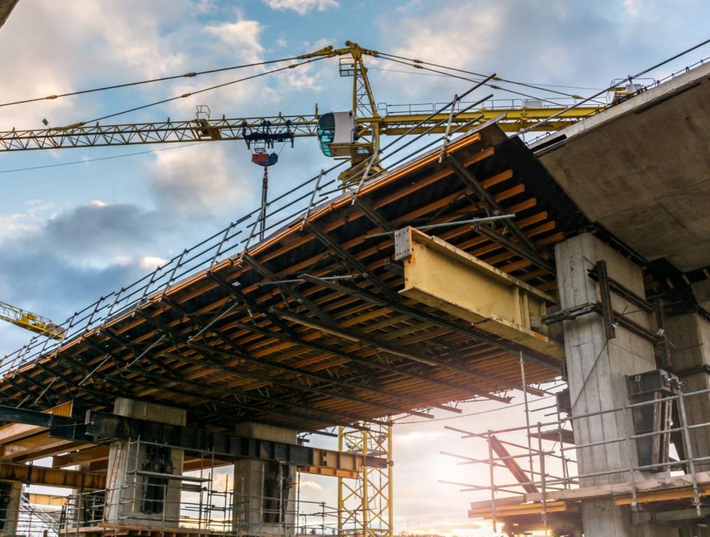 Photo of a bridge under construction, like the one being built on the 215 beltway in Las Vegas, shows wooden beams being fitted by a crane.