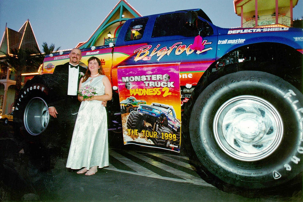 Two couples smiling in front of a monster truck that also has a sign that it promoting the show.