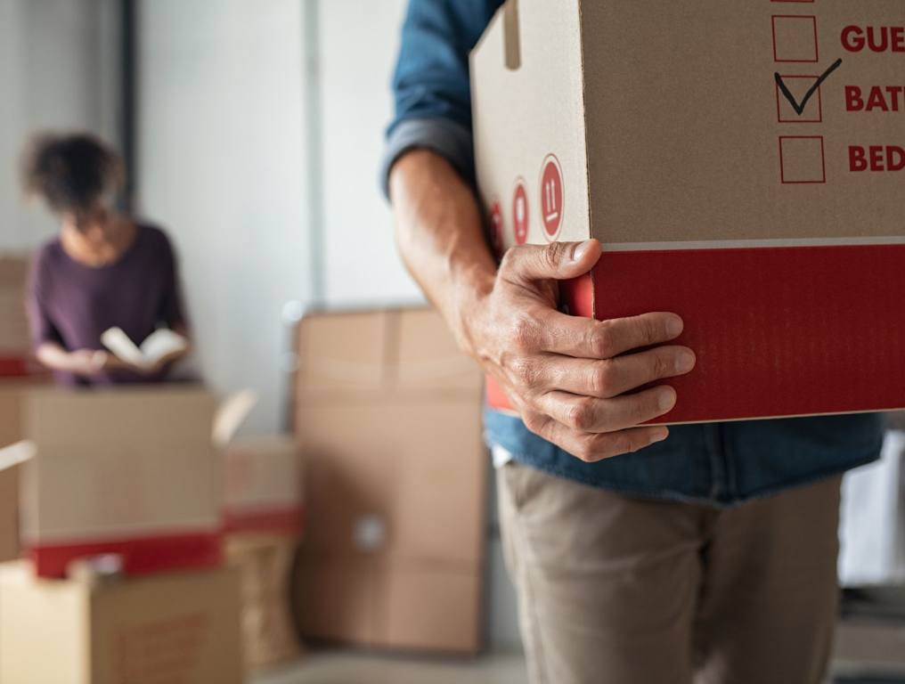 Close up of a moving box with a man's hands around it. There's a woman sitting on the couch in the background surrounded by more boxes. Concept: moving to Vegas