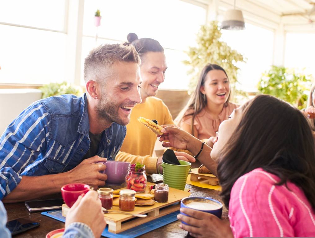 A group of friends sits around a brunch table in a cafe sharing food and laughing. Concept: Broken Yolk Cafe