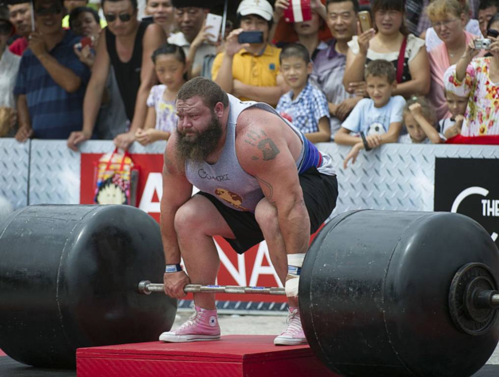 A large man with a long beard and short hair is crouched down in preparation to pick up a large barbell in a strong man competition. Concept: USA Strongman Championships