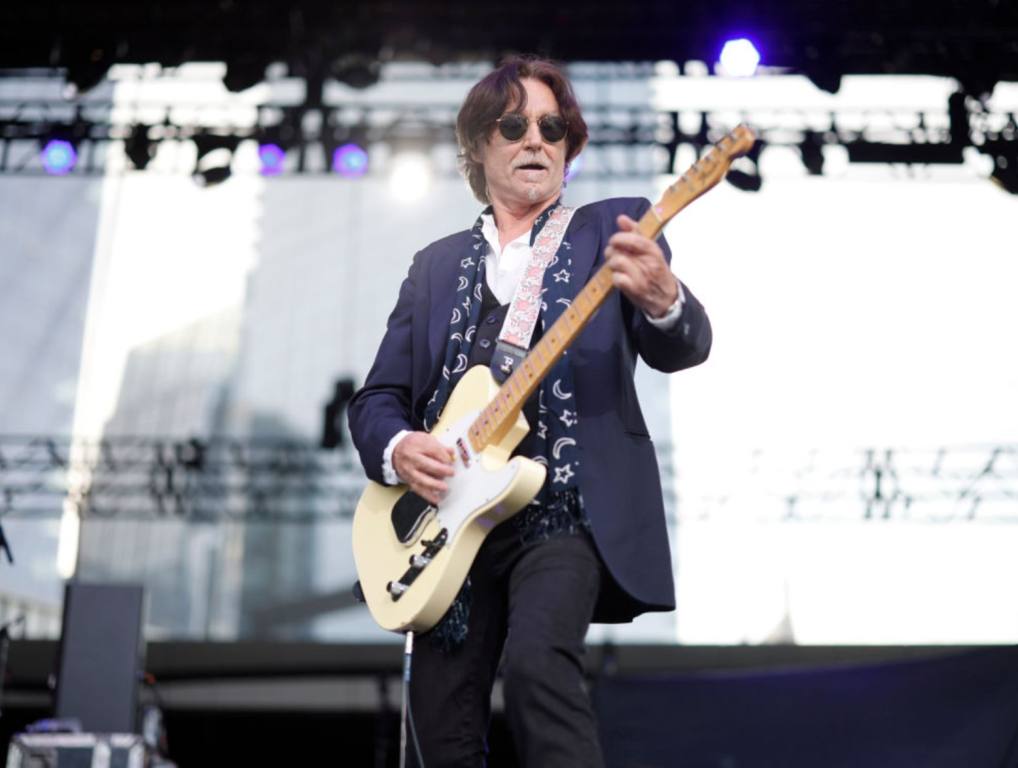 Singer songwriter John Waite performs onstage in a blue jacket and playing a white electric guitar.