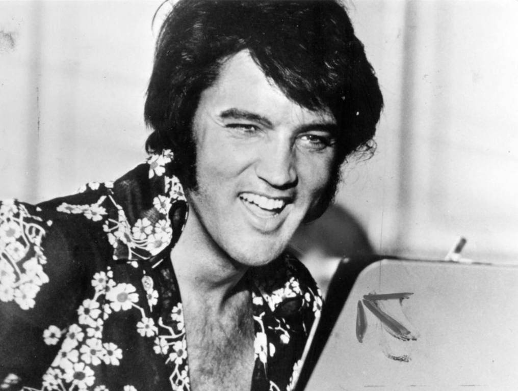 Black and white photo of Elvis Presley wearing a black button up shirt with white flowers. He is looking off to the side and smiling. Concept: Ghost of Elvis