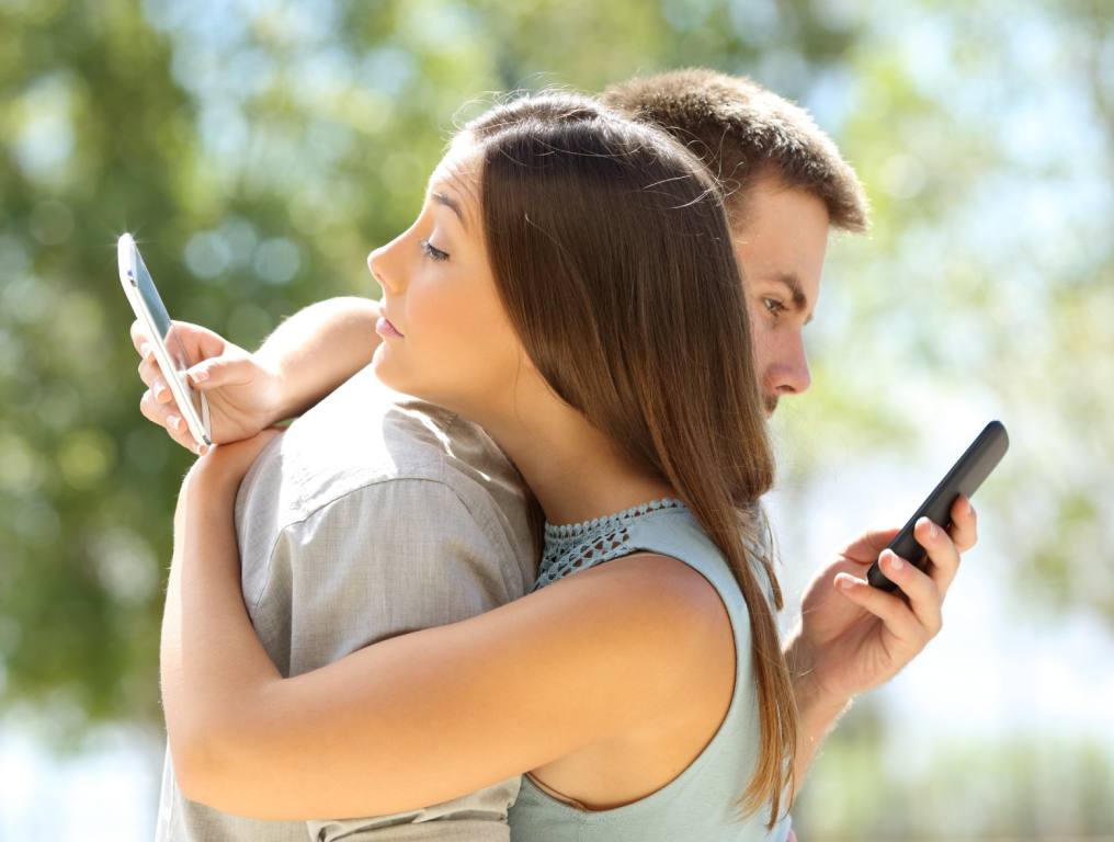 Profile picture of a man and woman hugging with each of them looking at their cell phones behind the back of the other. Concept: Las Vegas Dating Scene