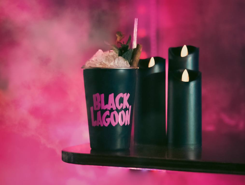 A black plastic drink cup with the words "Black Lagoon" spelled out in pink sits on a black tray with black LED candles. The background is pink fog from a smoke machine.