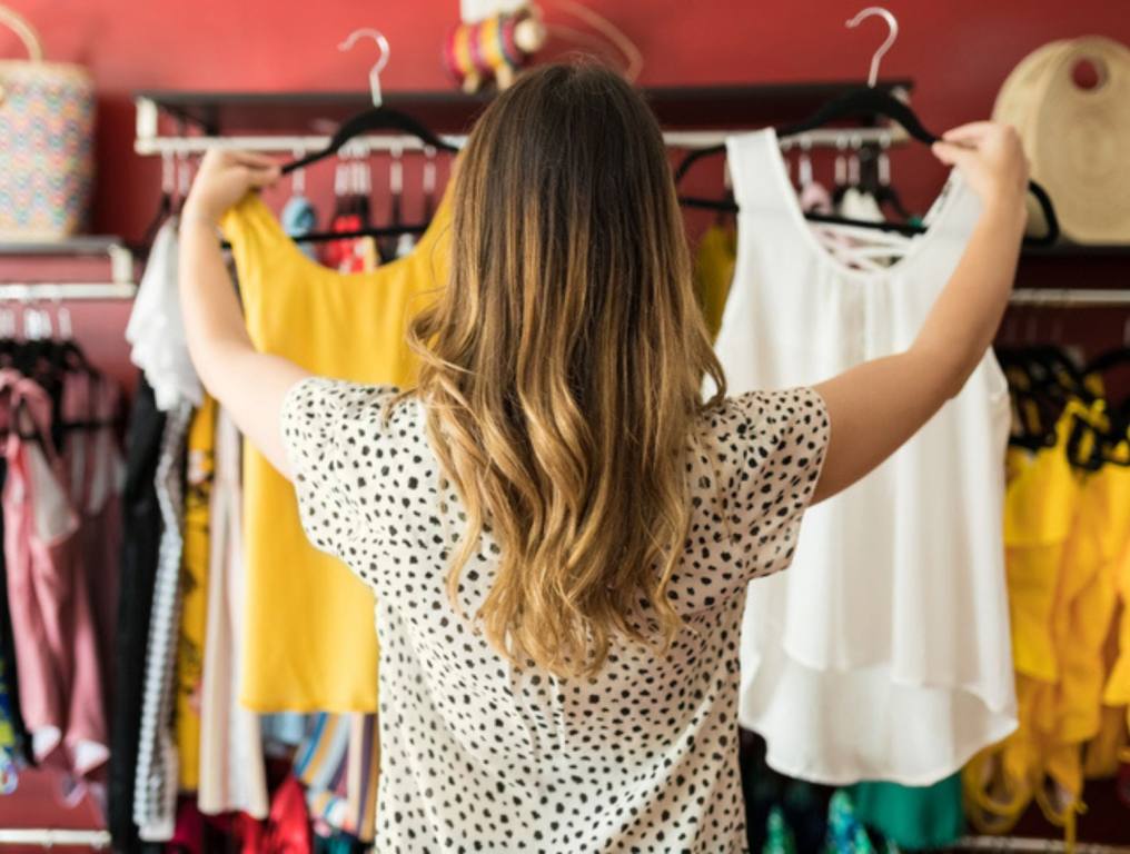 Rear view of young female customer with long brown hair choosing between tops at shopping mall