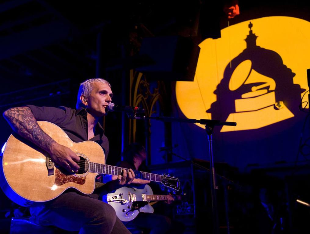 Blonde male lead singer of Everclear on stage holds an acoustic guitar and sings into a microphone as he sits in front of a lighted backdrop.