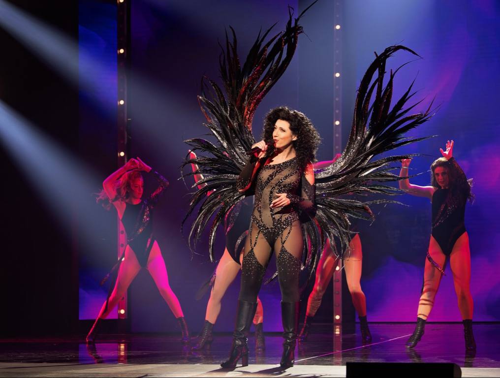 An actress portrays superstar Cher in an iconic black costume with black angel wings. She's on stage singing during The Cher Show.