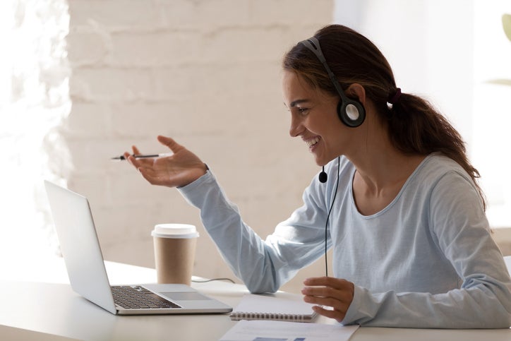 Side view head shot smiling mixed race lady freelancer wearing headset, communicating with client via video computer call. Millennial pleasant professional female tutor giving online language class.