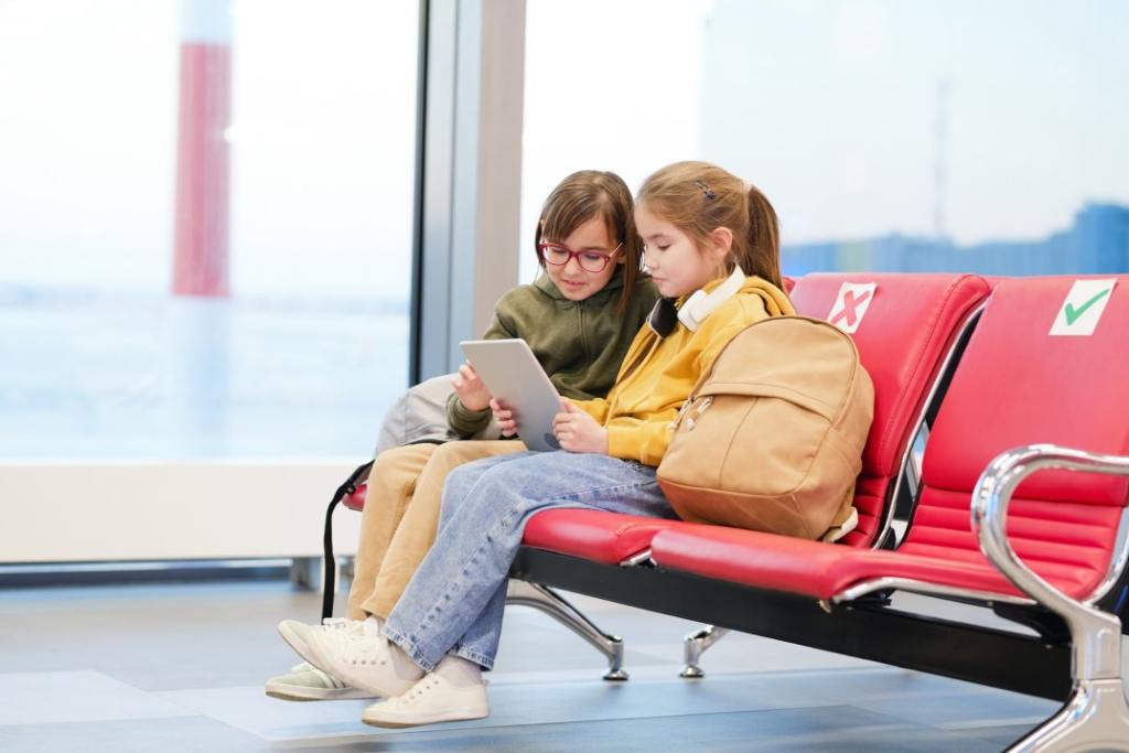 Two cute little girls in casualwear using digital tablet while sitting in lounge of modern airport and watching cartoons or movie for children.