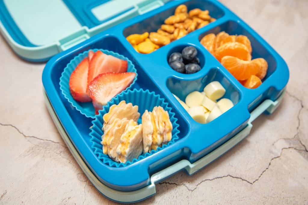 A blue bento box filled with strawberries, goldfish crackers, banana slices and oranges.