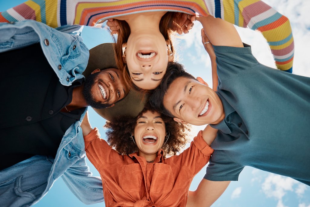 Group of diverse friends standing in a huddle against a blue sky. Multi-racial friends standing together arms around each other and smiling while looking down at the camera.