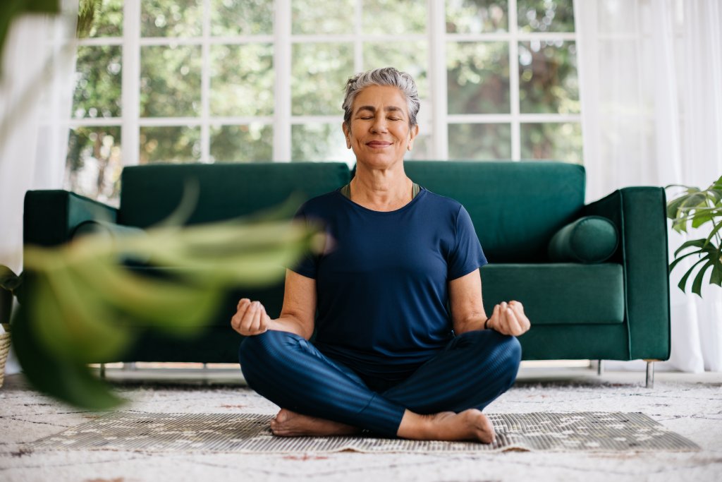 Senior woman meditating in lotus position at home, sitting on the floor in navy blue fitness clothing. She is sitting in front of a green modern sofa.