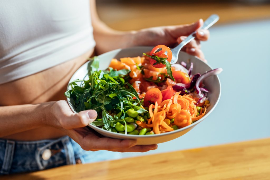 Close up of a bowl of fresh veggies being held by a woman in a white crop top. She is holding a fork with food on it.