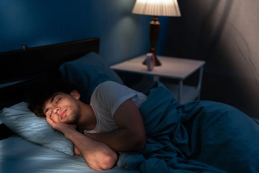 A young dark-haired man is sleeping in bed. A lamp is turned on behind him on the nightstand and the light from the window is shining on his face.