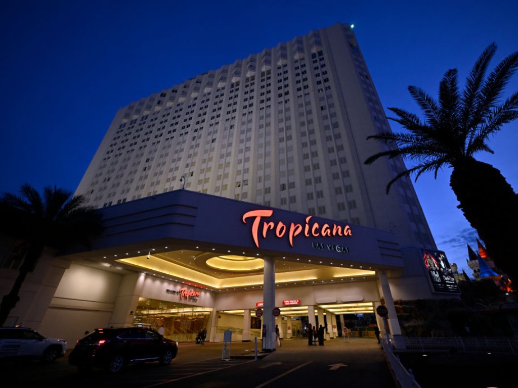 An exterior view shows the Tropicana Las Vegas at dusk. The hotel is white and the lettering is red. There is a palm tree to the right.