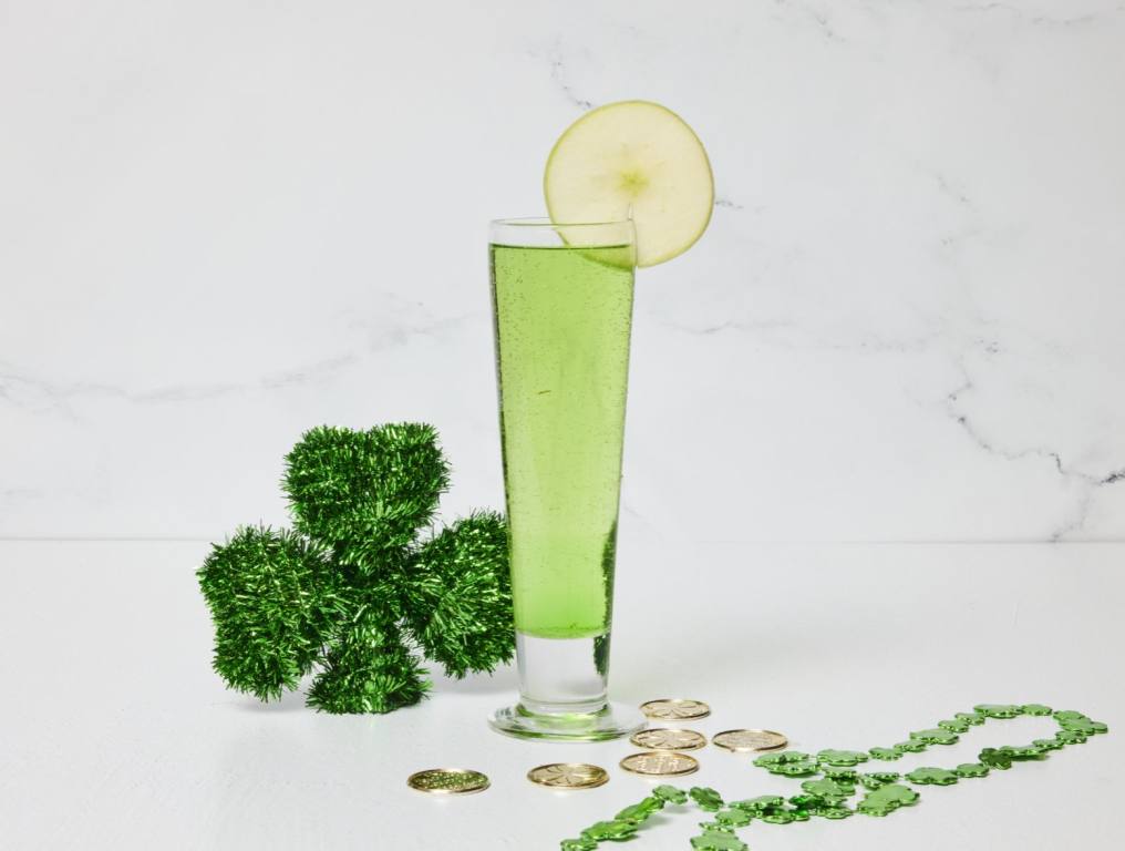 A green apple mimosa stands in a tall beer glass on a white table. It is garnished with a thin apple slice and surrounded by St. Patrick's Day decorations.