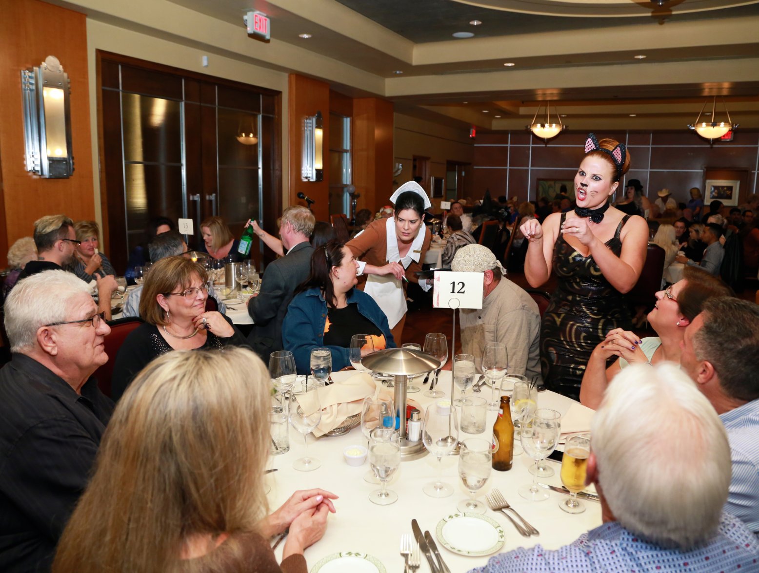 A group of people are sitting around a dining table at Lawry's Prime Rib. They are all smiling and looking at the server. Who is dressed in cat ears and whiskers. She has her hands up and her mouth open, like she is telling a story. The event is the St. Patrick's Day murder mystery dinner in Las Vegas, which many families enjoy for the holiday.
