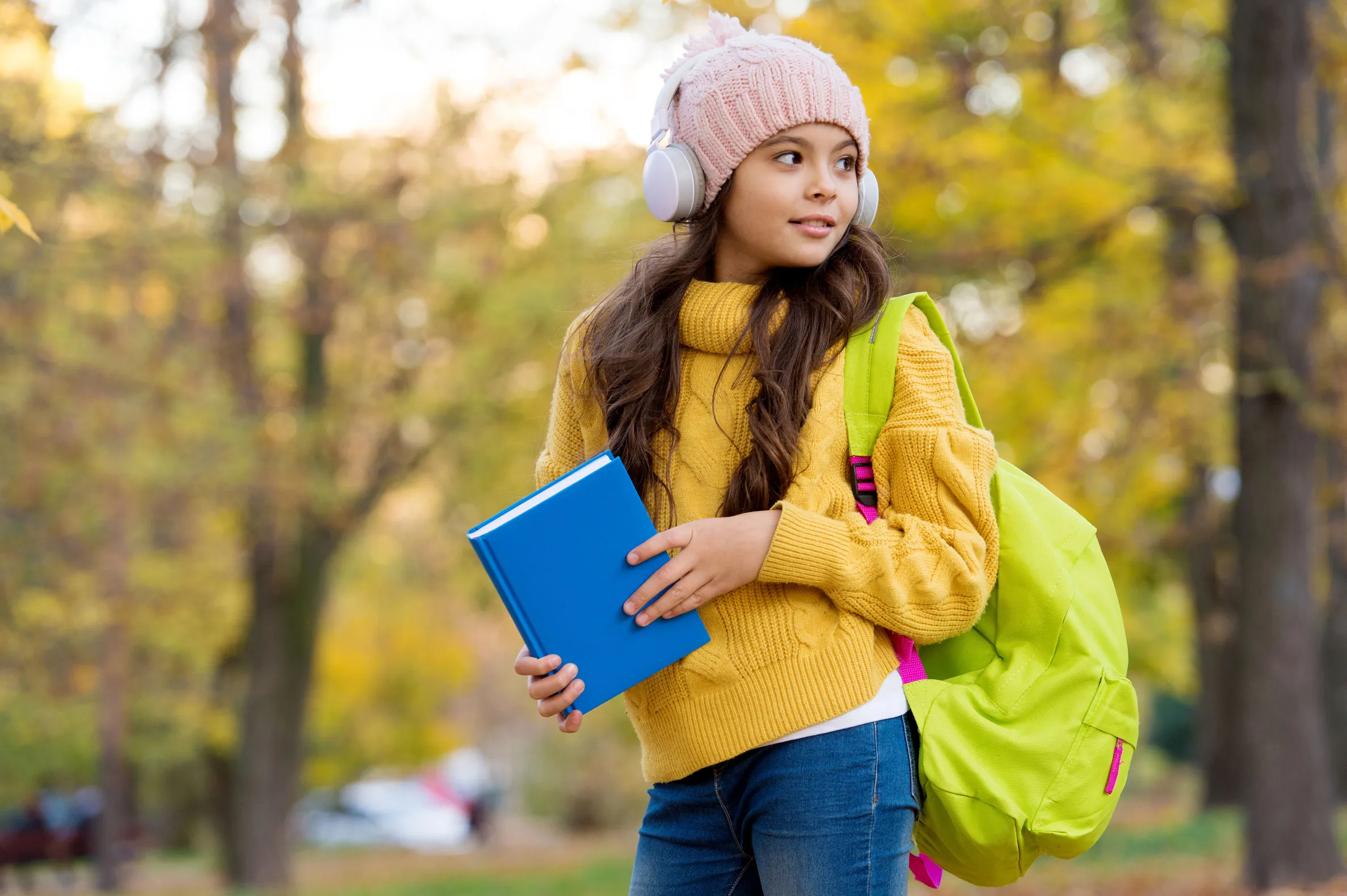 A young girl with brown hair in a yellow sweater holding a blue book. She is tanding in front of trees.
