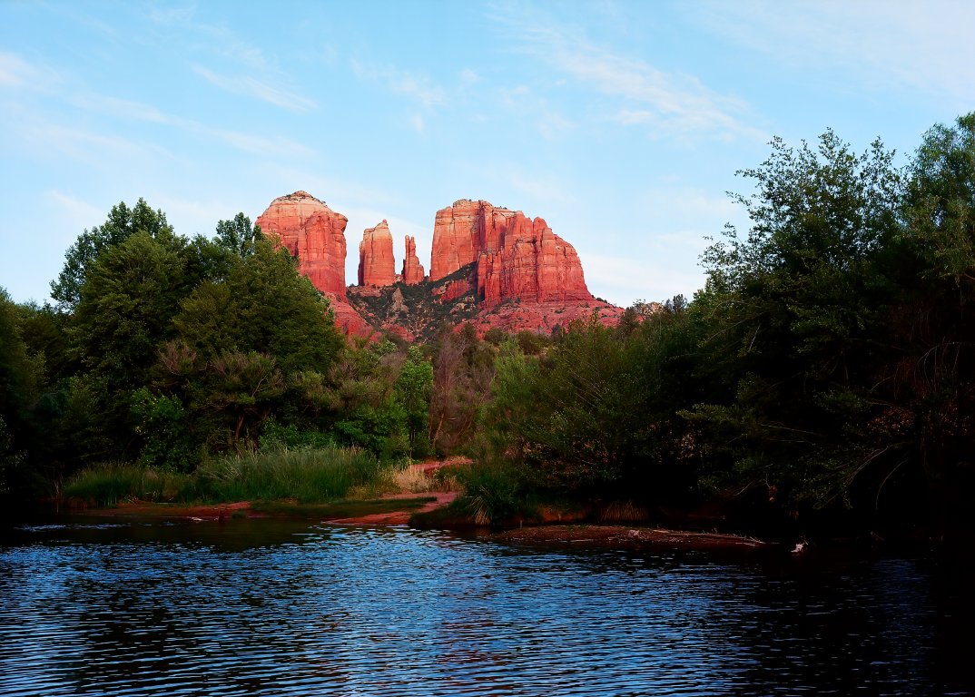 A long shot of Cathedral Rock in Sedona, Arizona shows red and cream sandstone layered in hoodoo formation. There is a river and trees in the foreground.