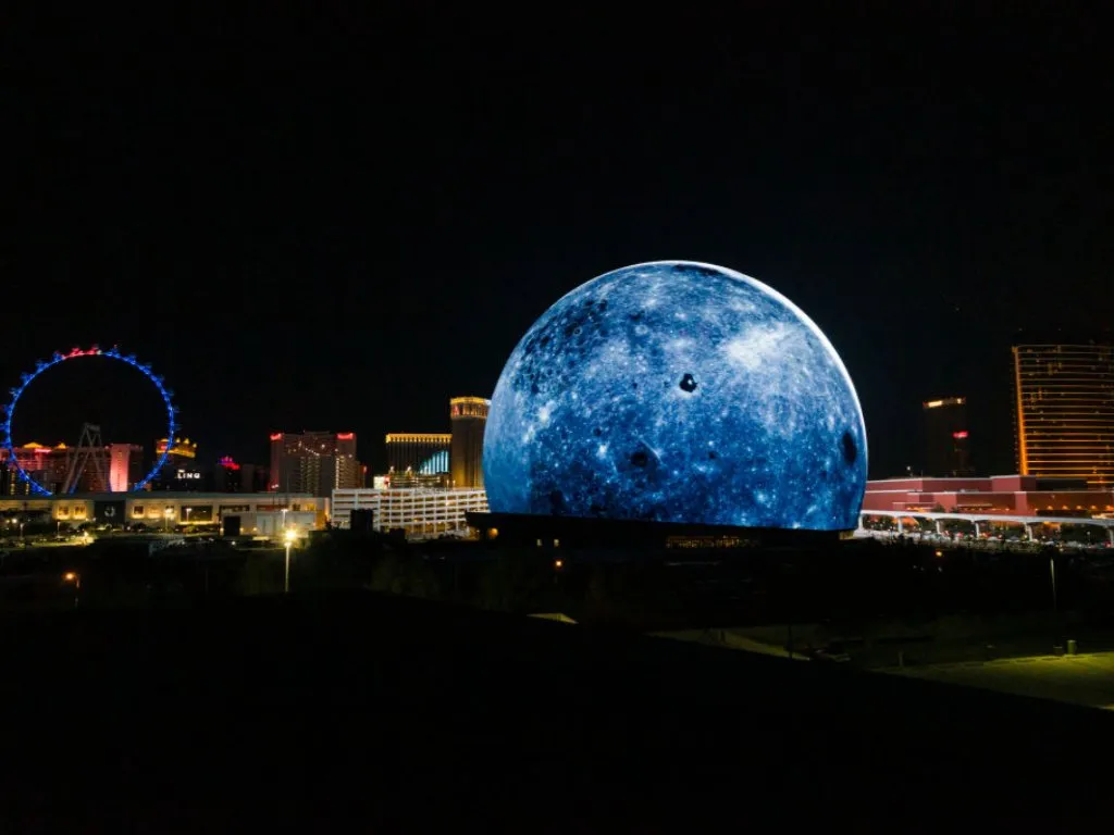 The Las Vegas Sphere is lit up to look like the planet Earth. It is in the foreground with the Las Vegas Strip in the background. The sky is dark and the casinos are lit up.