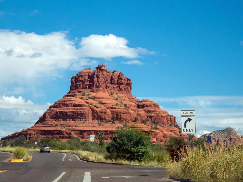 A red sandstone mountain lies at the end of a winding road. Blue skies and white clouds are above it and there are trees on the shoulder.