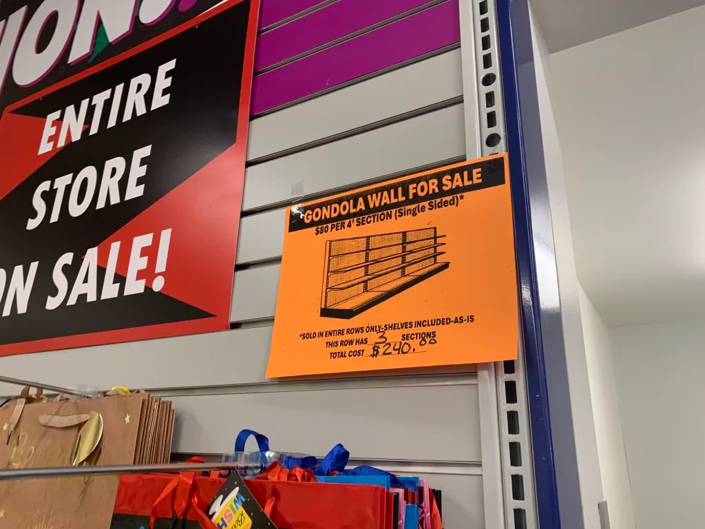 A retail gondola wall is displaying gift bags. There is an orange printed sign indicating the wall itself was also for sale.