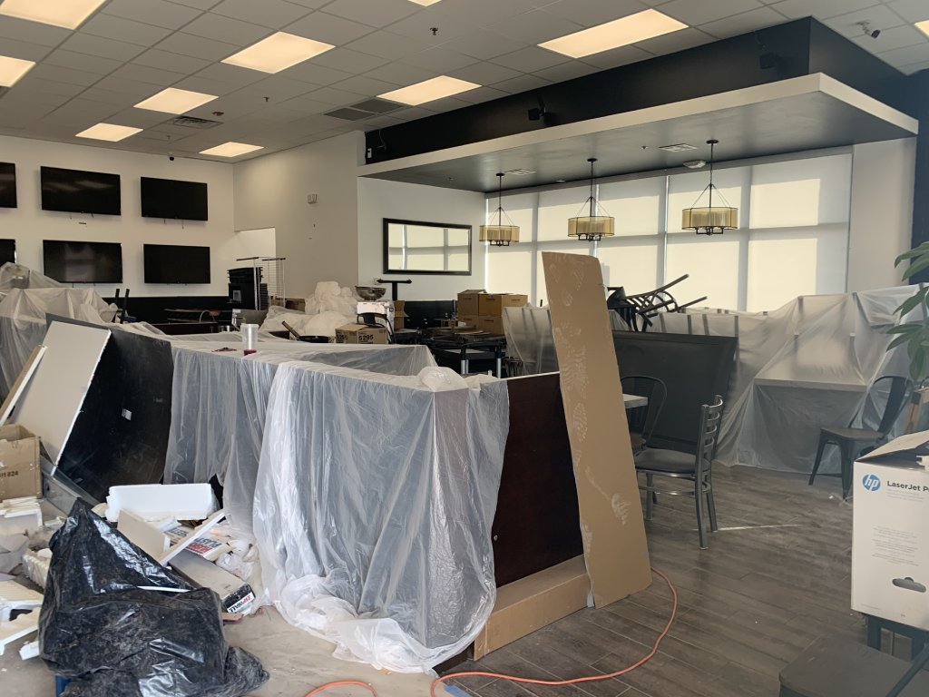 The interior of a restaurant under redesign has dining booths under painting plastic. In the background are six tvs and hanging lamps.