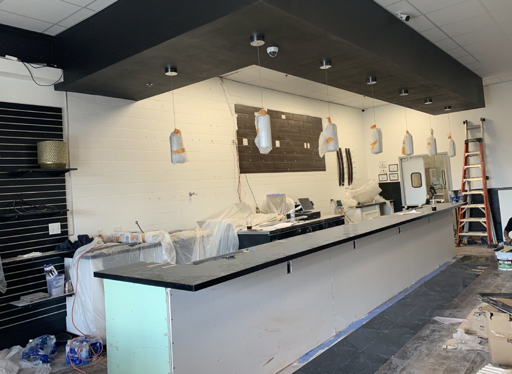 The interior of a restaurant under construction shows a white bar with dark brown countertops and scones hanging. Much of the room is covered in painting plastic and tape.