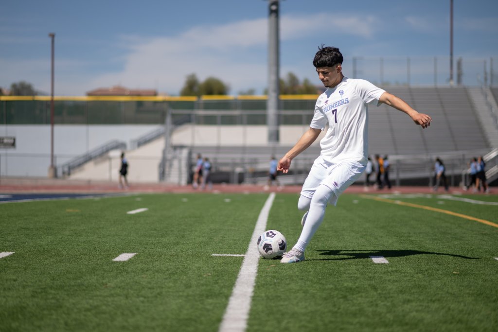 A young latino boy in a white uniform kicks a soccer boy on a high school field.