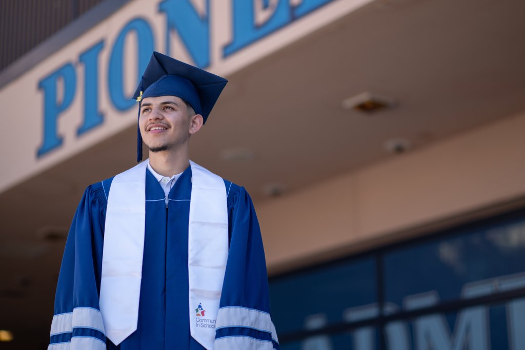 A latino boy in a blue cap and gown is standing in front of his school. He is smiling and looking out as if contemplating his future.