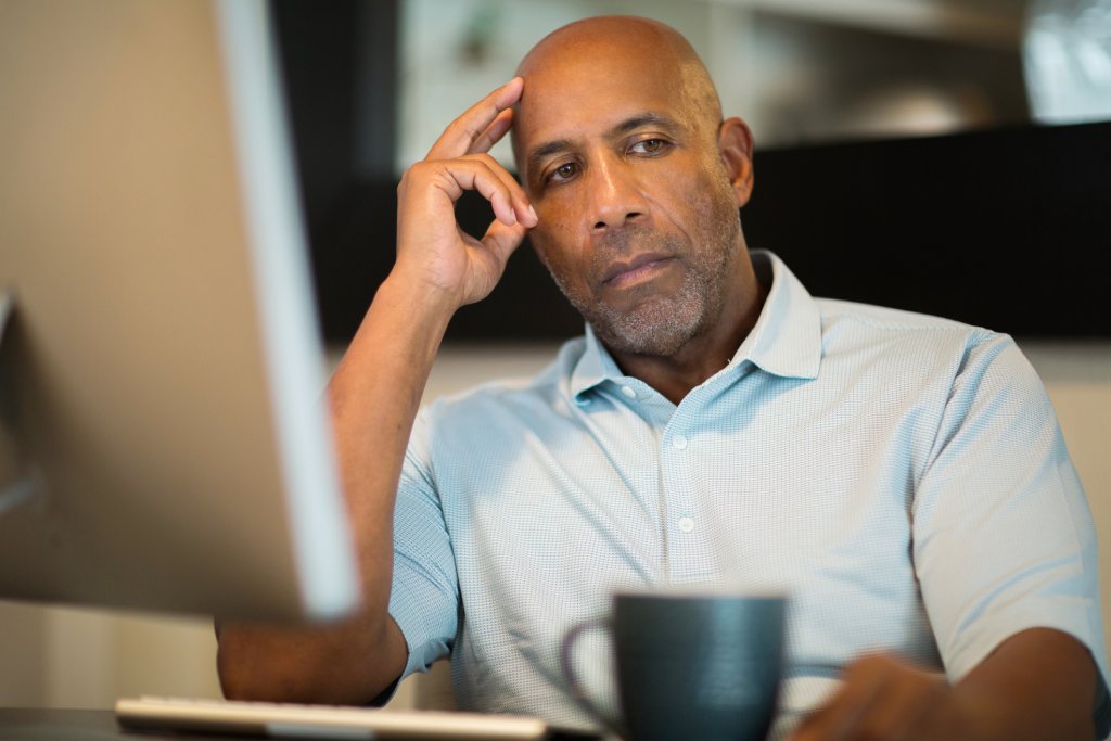 Mature black man looks worried as he sits in front of his open laptop with his fingers touching his temple and his gaze off to the side.